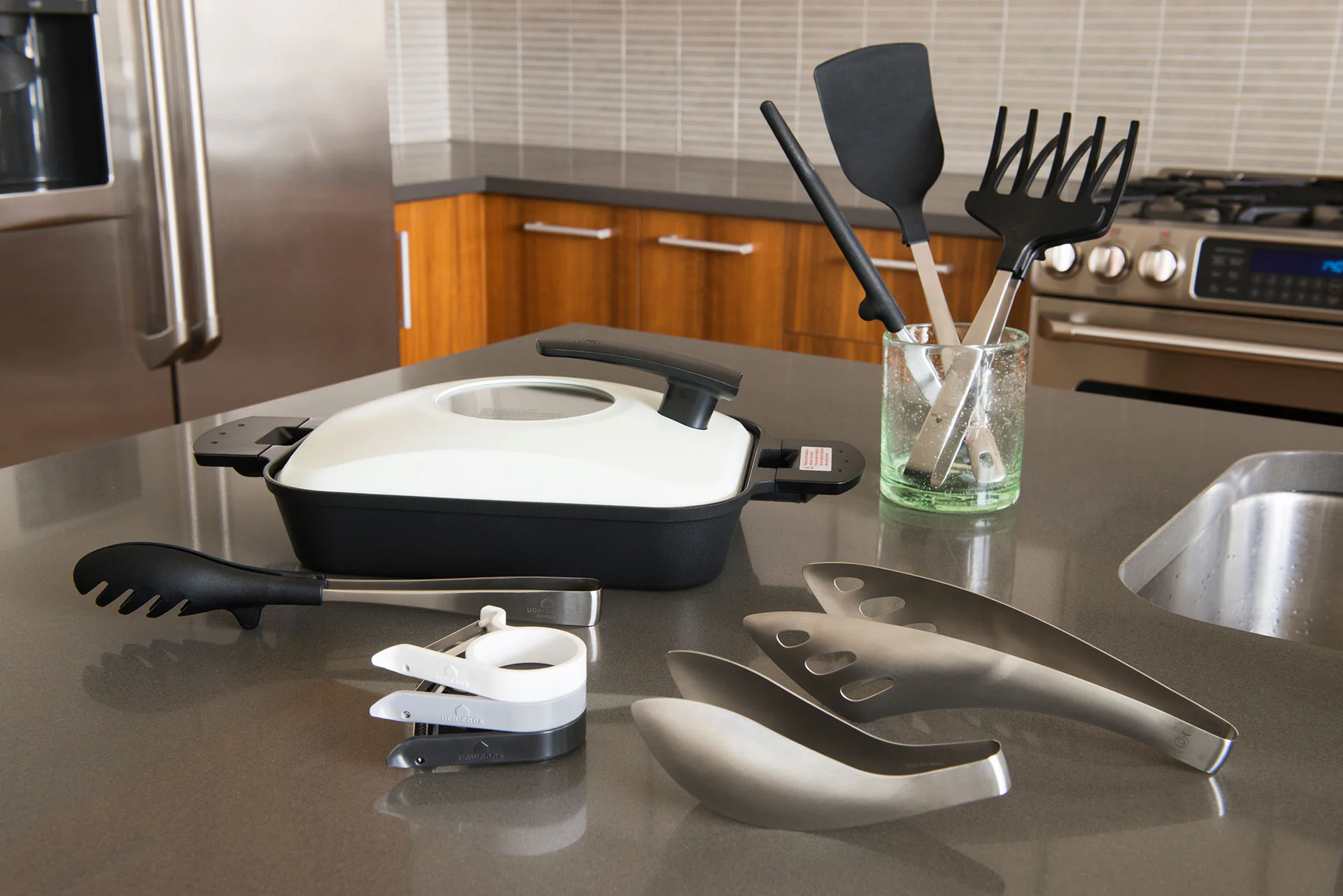 Uchicook-Pfanne mit weißem Deckel und Küchenhelfern auf einer Arbeitsplatte.